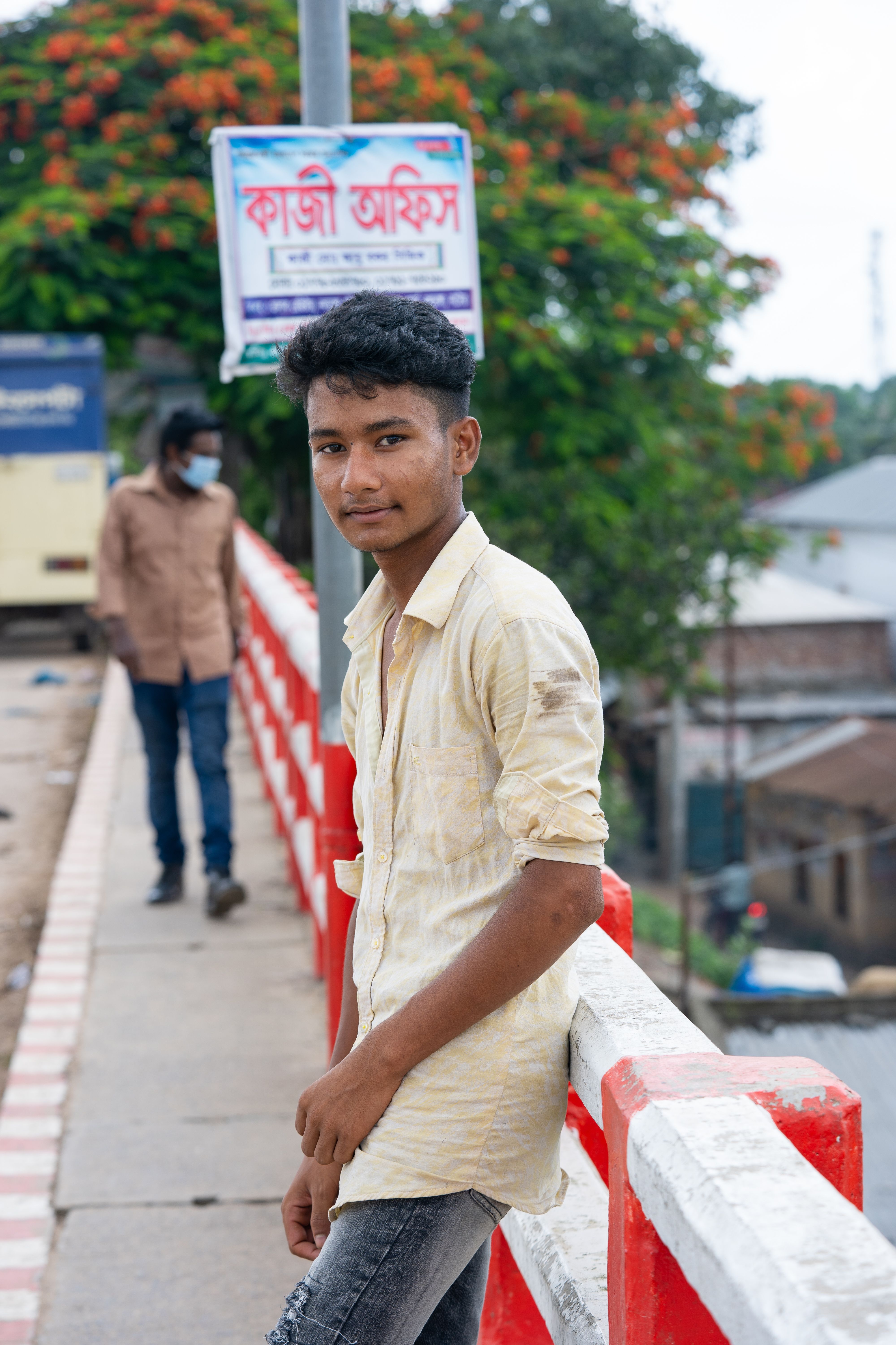 A young man in a yellow shirt leans on a red and white railing in Bangladesh, posing outdoors.