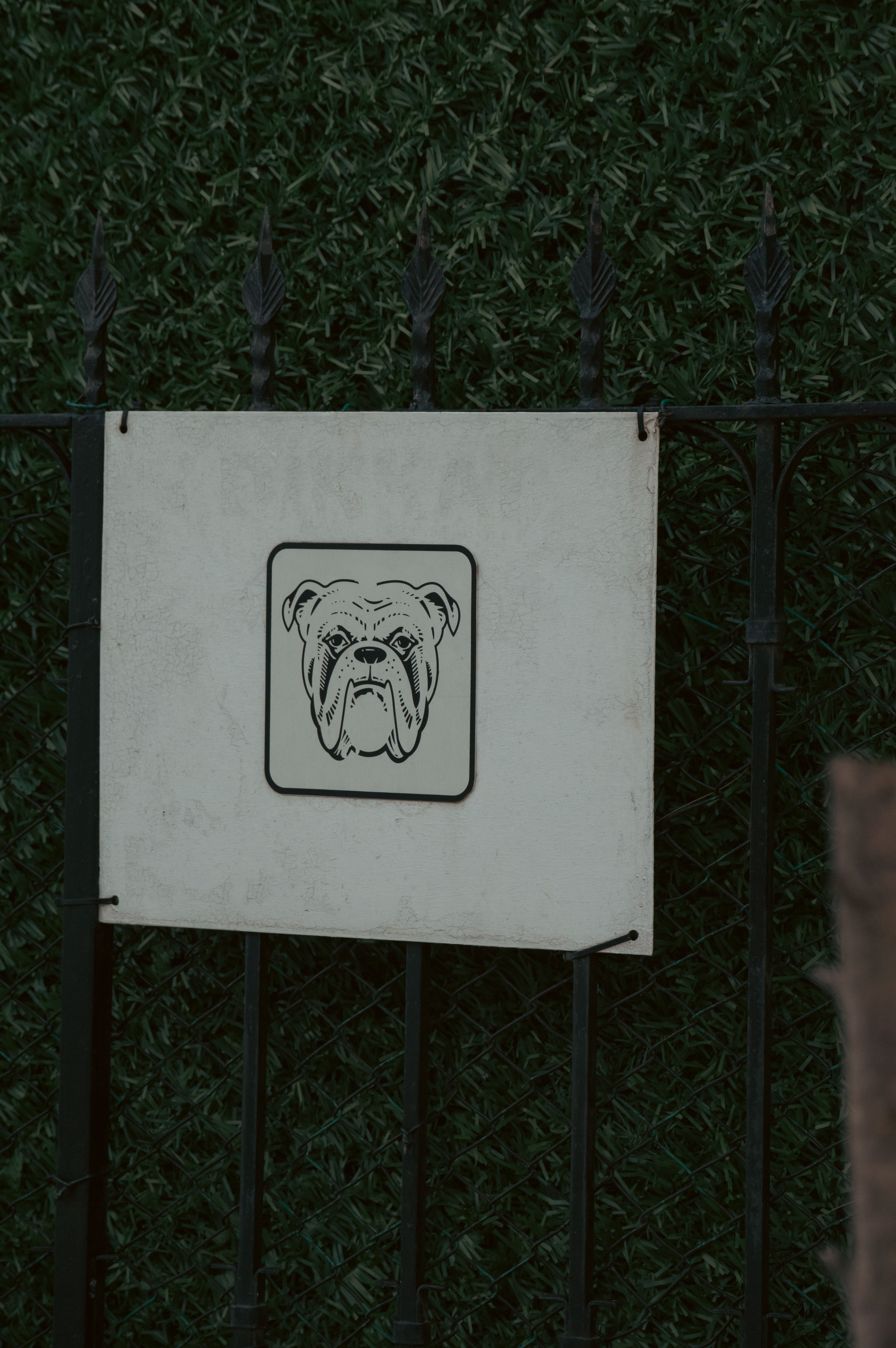 Illustrated bulldog warning sign on iron fence with greenery background.