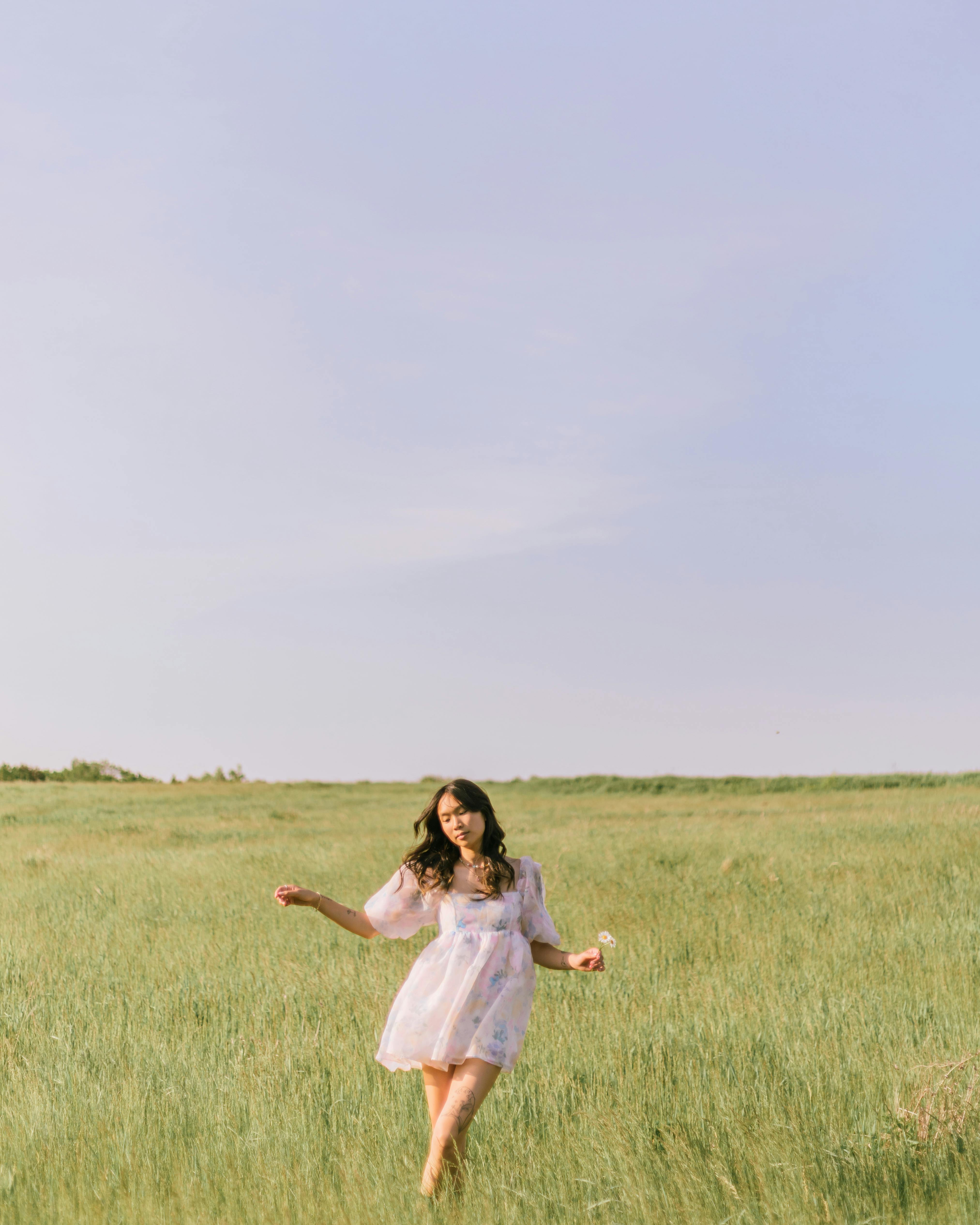 A woman in a white dress walks through a grassy field under a clear sky, evoking a serene summer mood.