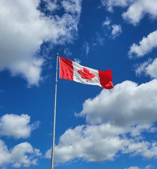 A low-angle view of the Canadian flag waving on a flagpole against a bright blue sky with clouds.