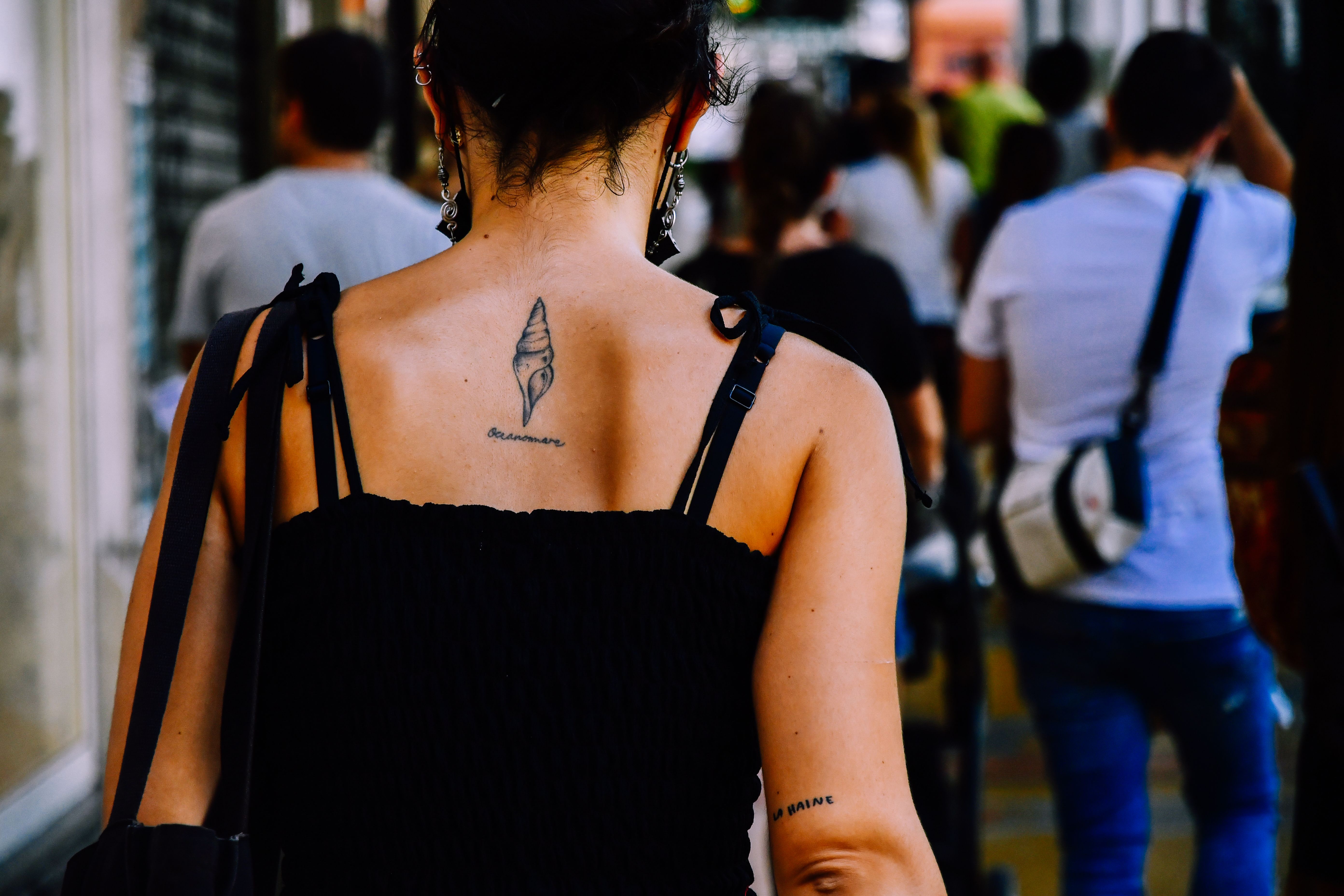 Back view of tattooed woman in black tank top walking down a bustling city street.
