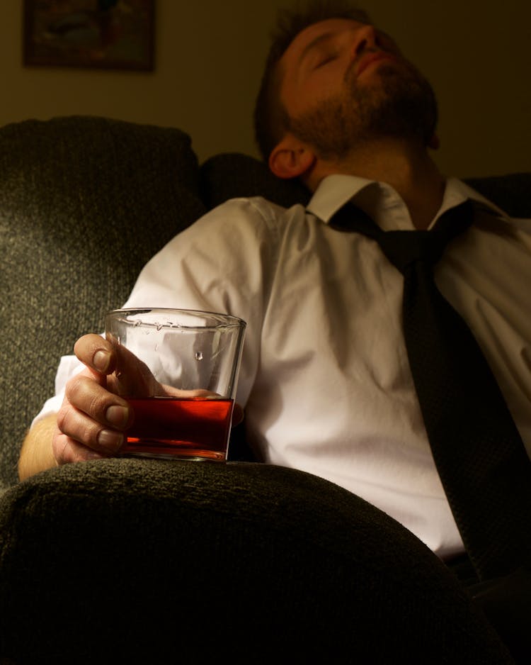 A Tired Man In White Long Sleeves Resting On A Couch While Holding A Glass Of Whisky  