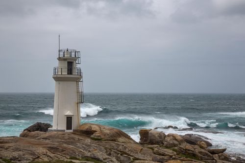 Free White lighthouse on rocky coastline in Galicia, Spain amidst crashing ocean waves. Stock Photo