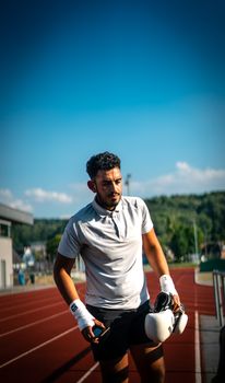 A young male boxer resting on a track holding boxing gloves with a clear blue sky overhead.