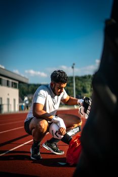 A male athlete adjusts his boxing gloves on a running track, preparing for training under a clear sky.