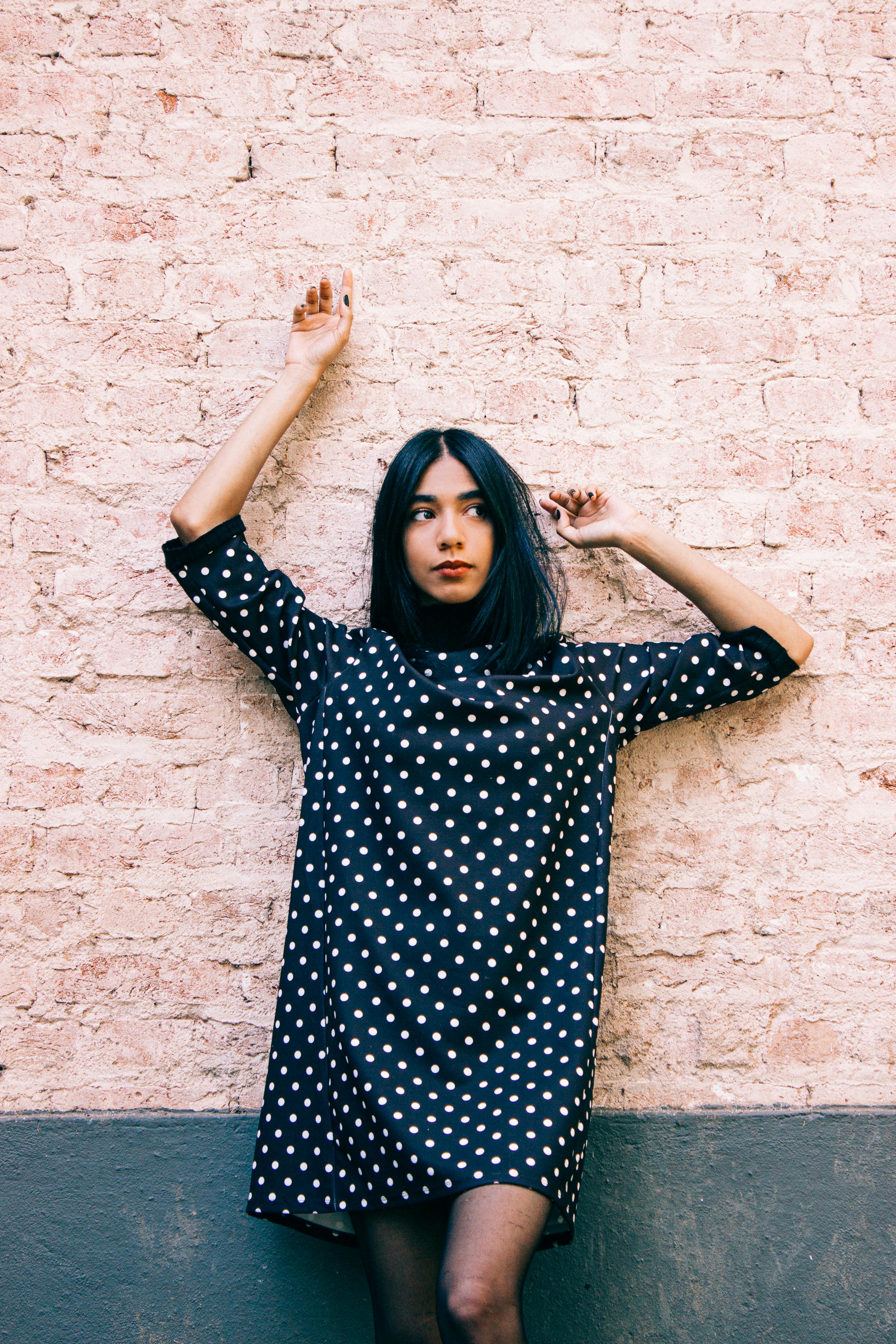 Woman in a polka dot dress posing against a brick wall in an outdoor setting.