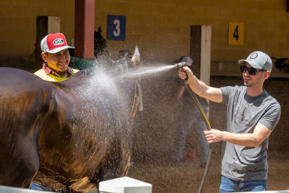 Two men using a water hose to wash a horse in an outdoor stable setting.