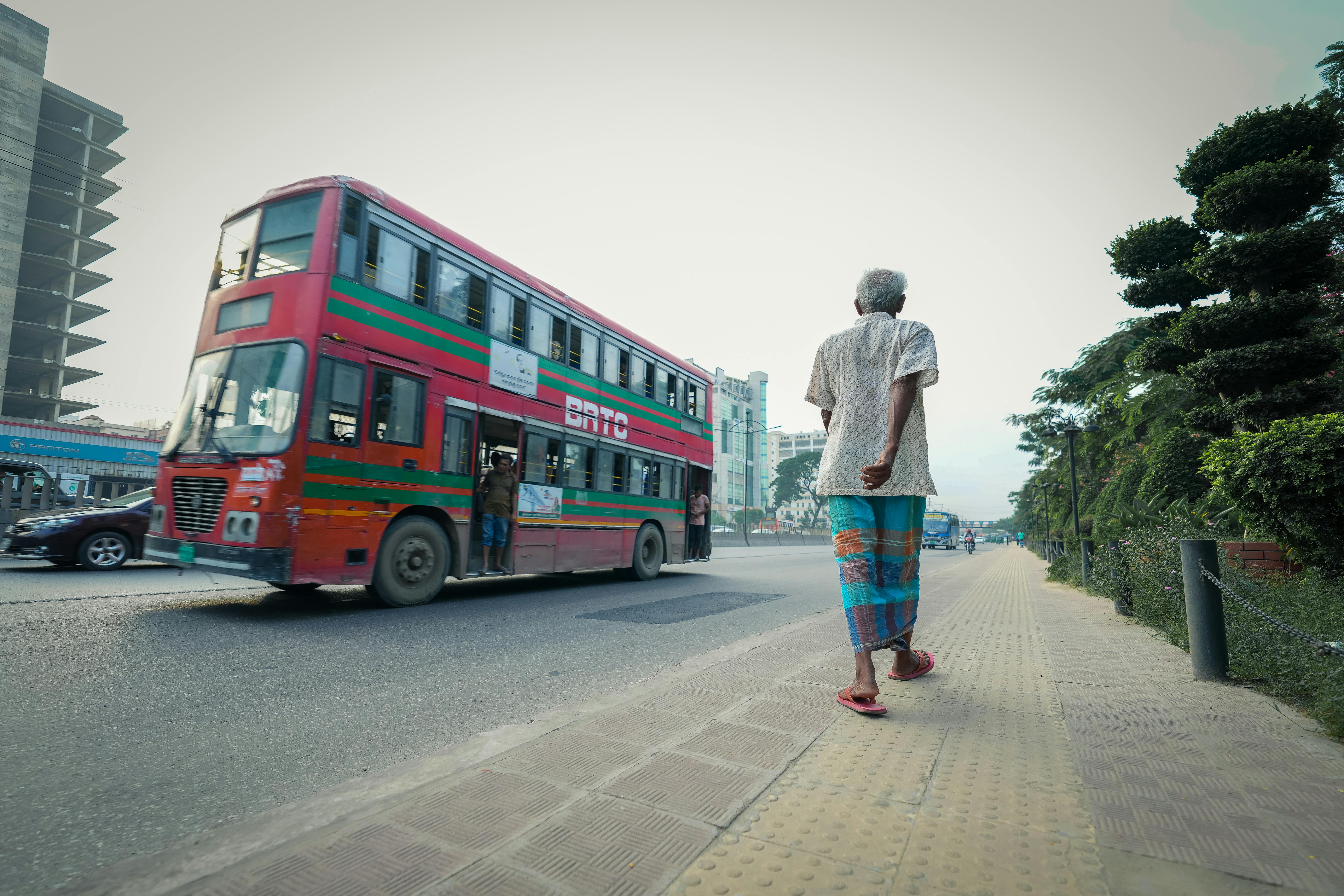 Free Elderly man walks along a busy city street with a vibrant double decker bus passing by on a sunny day. Stock Photo