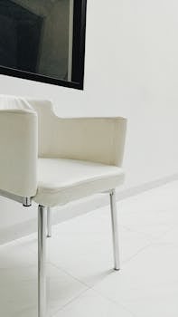 Modern white leather chair with chrome legs in a minimalist room, featuring a glass window.
