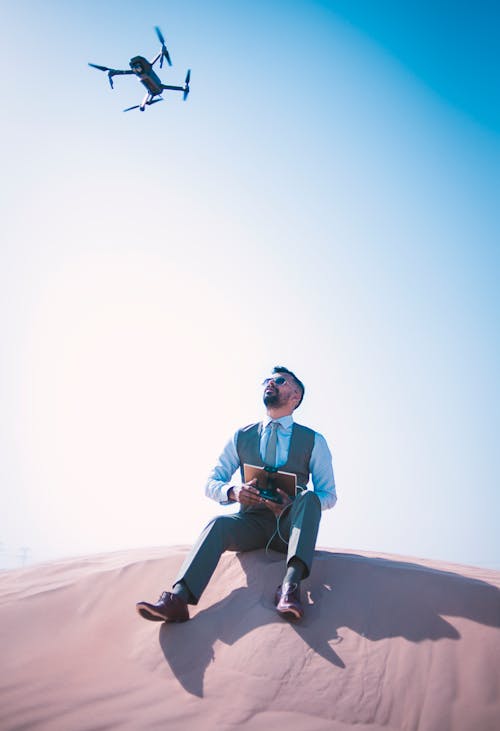 Man Sitting on Desert Ground Looking Up and Operating the Quadcopter