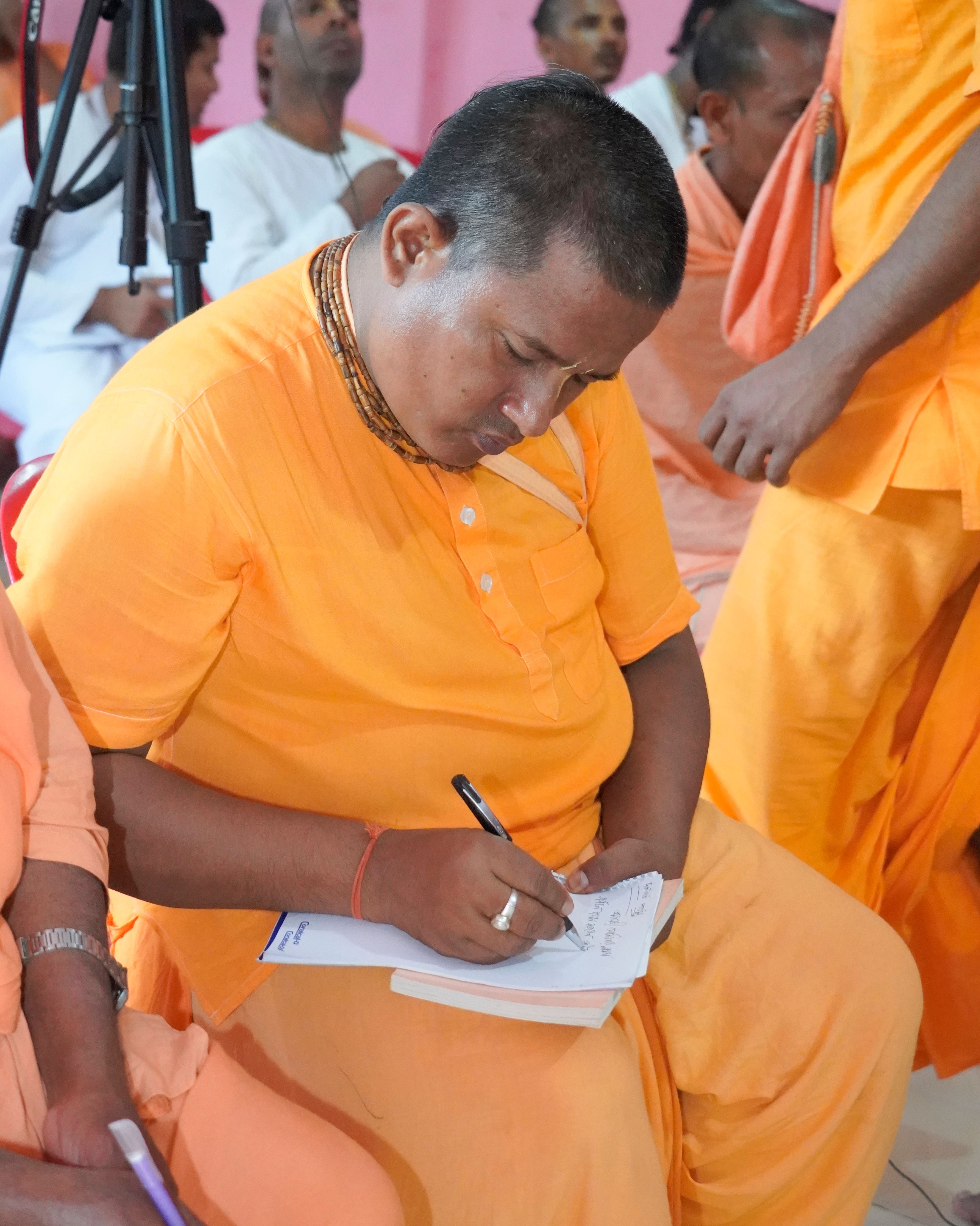 A South Asian man in traditional orange attire writes at a religious gathering, showcasing cultural practices.