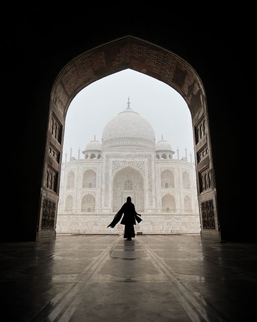 Silhouette of a woman in an arch framing the iconic Taj Mahal with foggy ambiance.