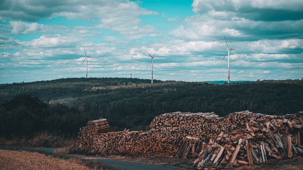 Wind turbines on a hill with stacked logs in the foreground under a cloudy sky.