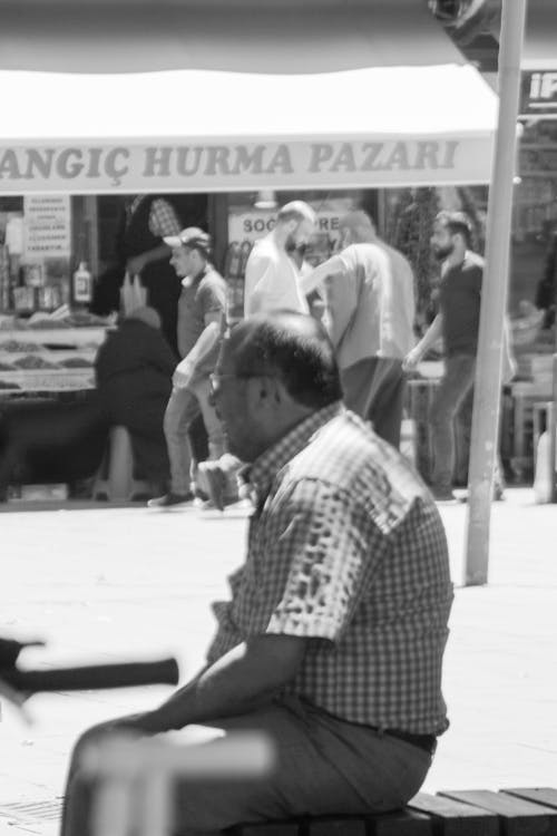 Grayscale Photo of Man Wearing a Checkered Shirt Sitting on a Bench