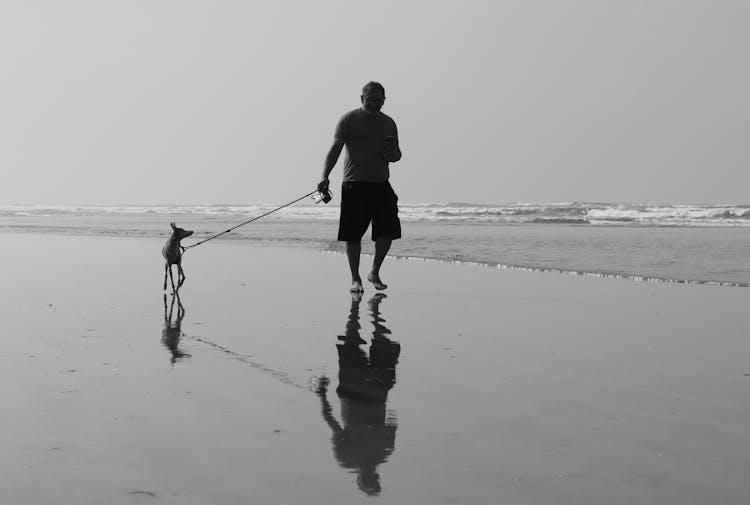 Black And White Photo Of Man Walking On Shore While Holding The Leash Of A Dog