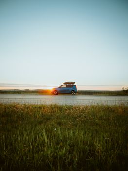 A scenic view of a blue car parked on a rural road at sunrise, with a serene landscape and a clear sky.