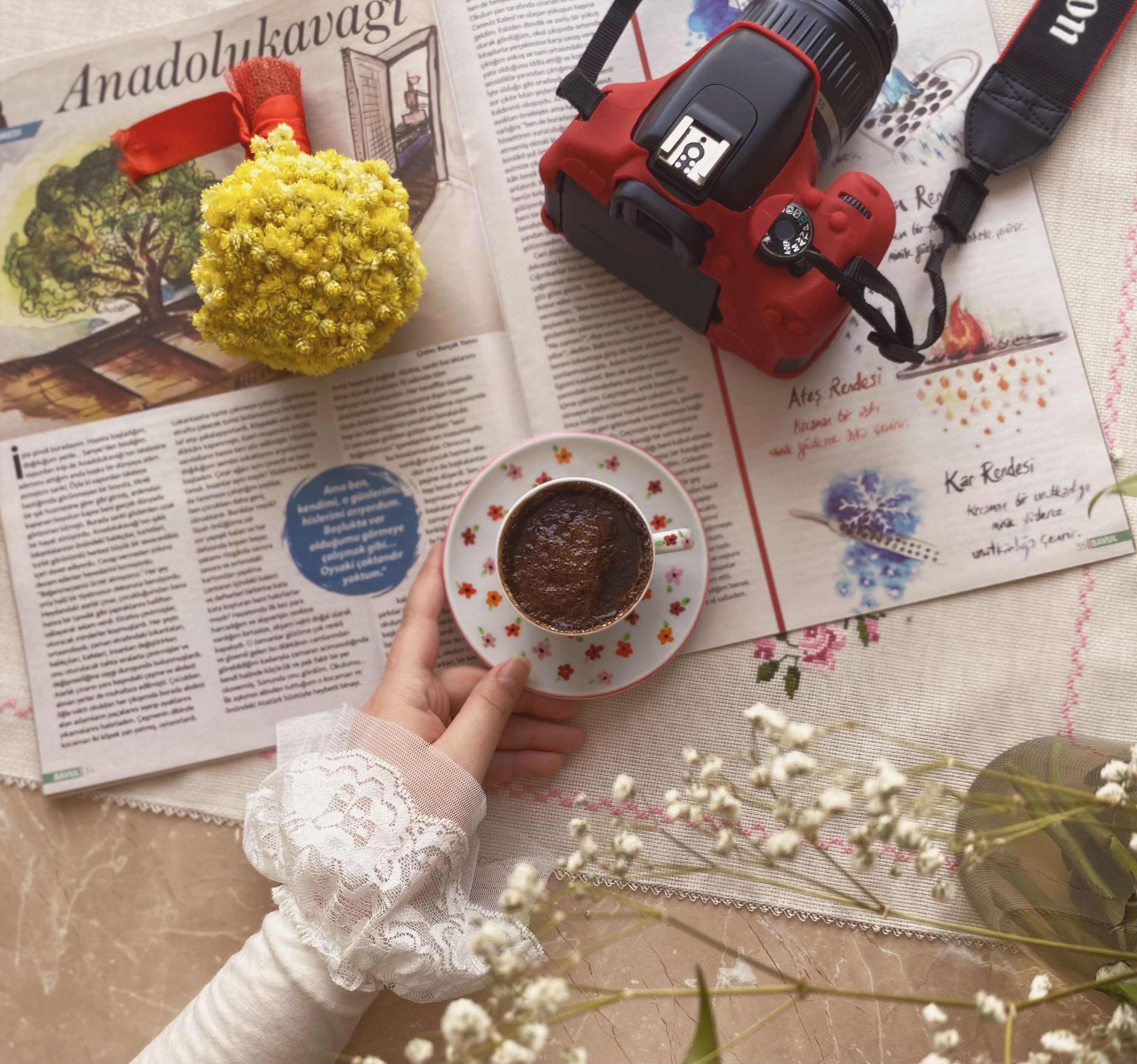 A cozy flat lay featuring a hand holding a cup of coffee, a camera, and a bouquet on a magazine.