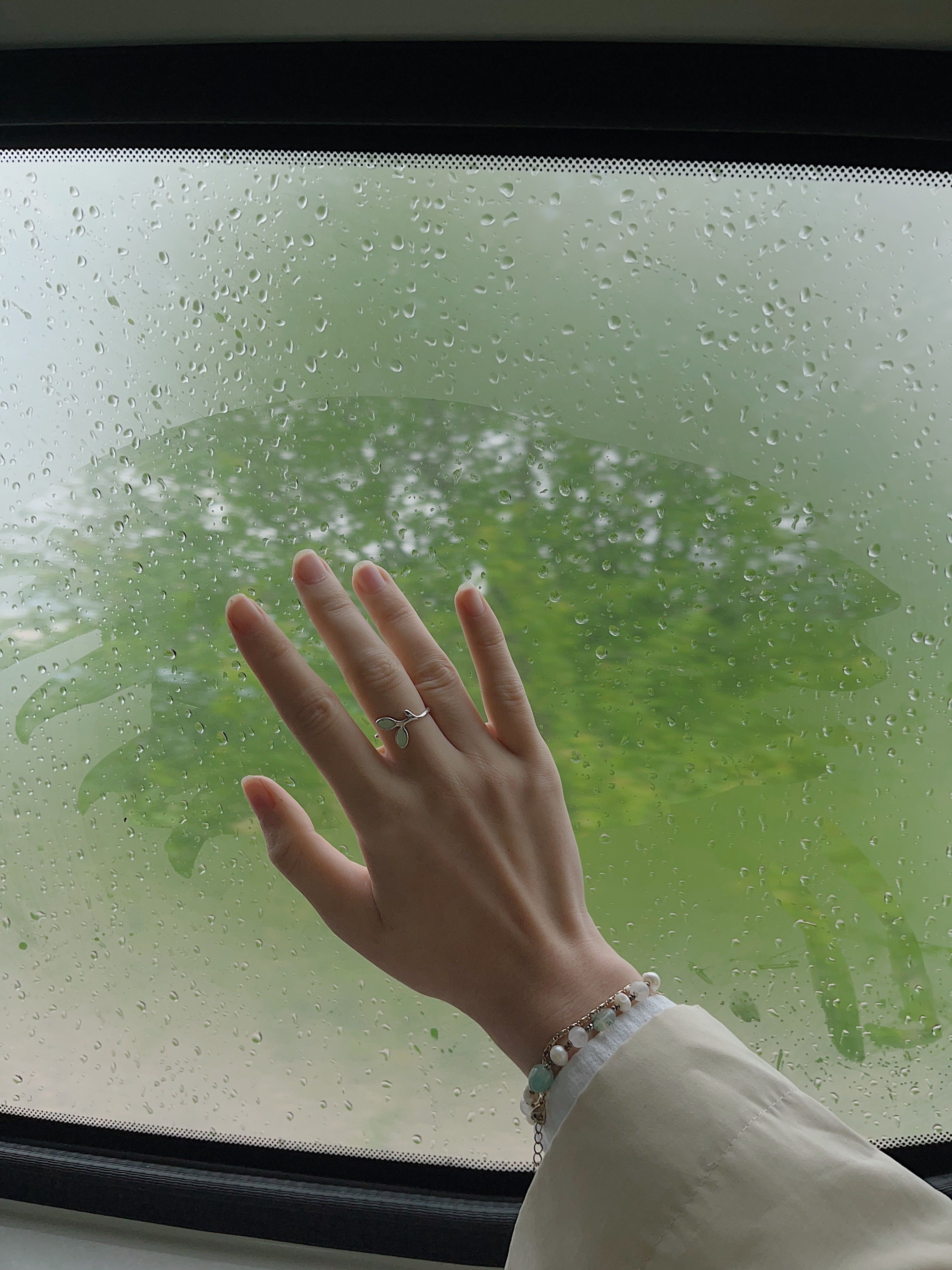 A close-up of a hand wiping raindrops off a car window, showcasing jewelry and moisture detail.