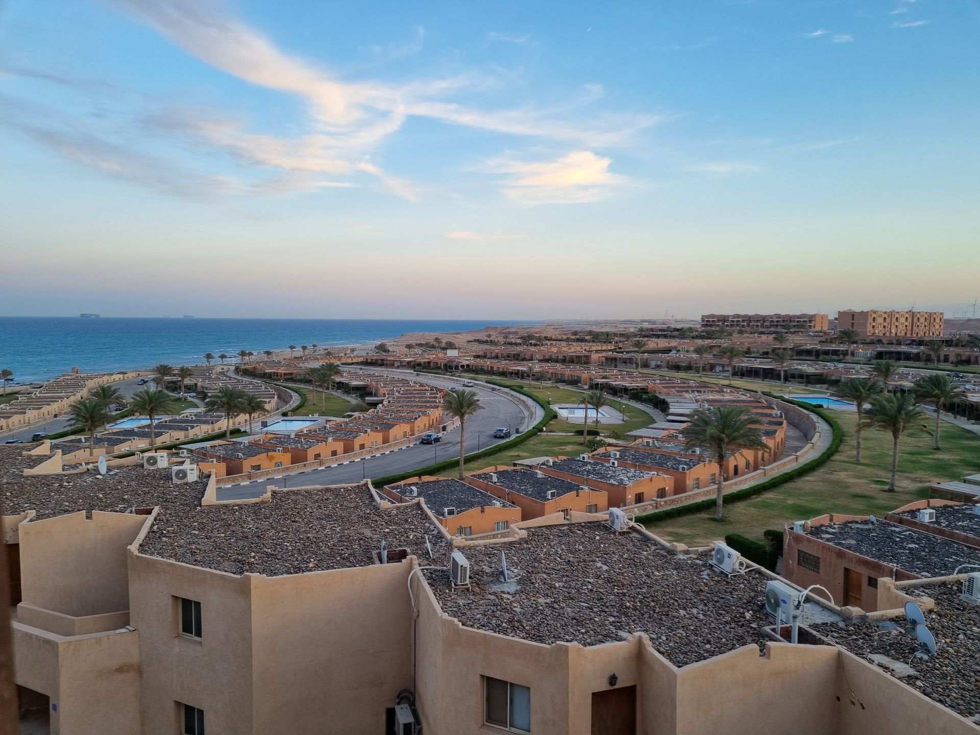 Aerial view of a residential area by the sea with palm trees and rooftops under a bright blue sky.