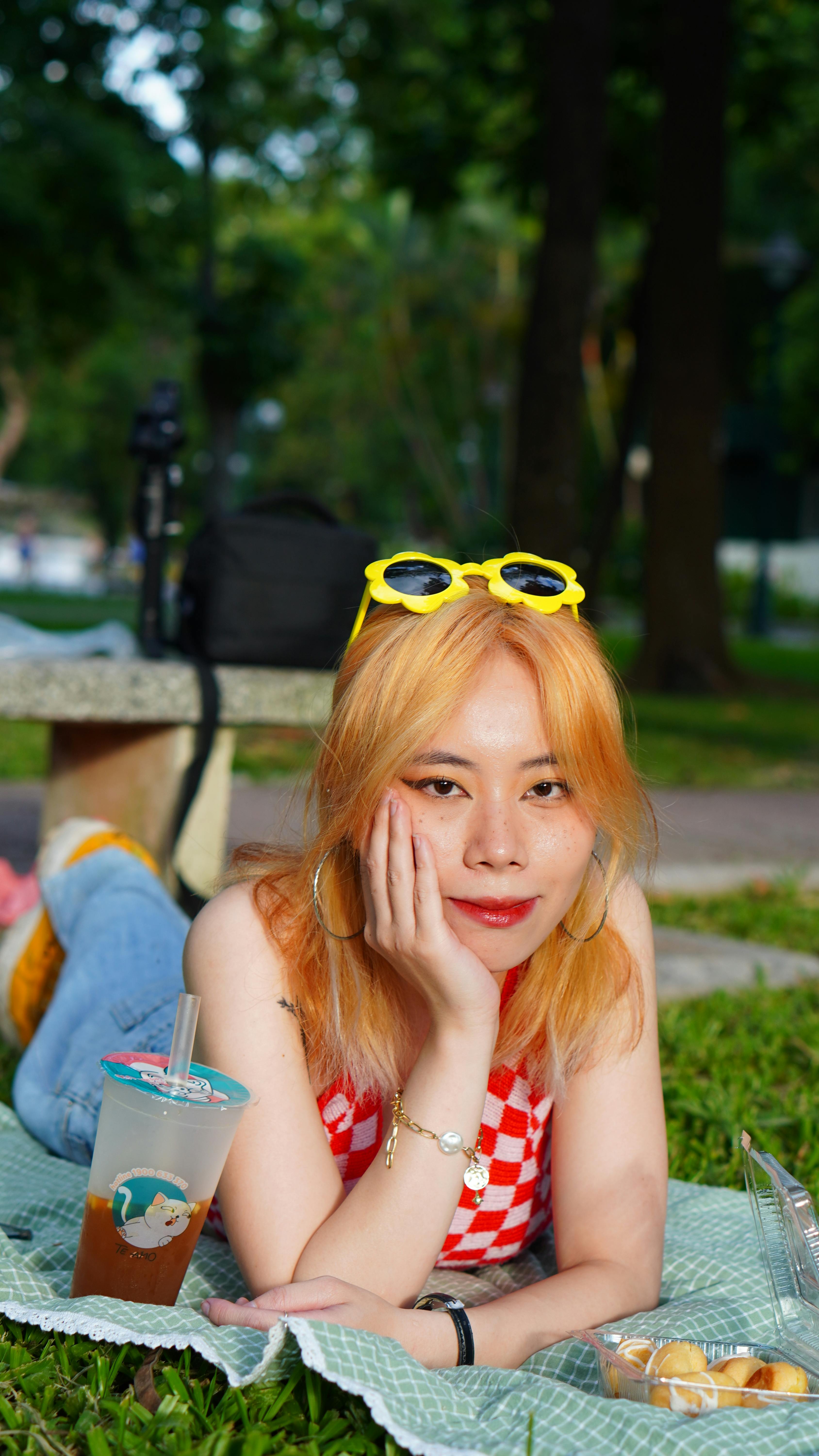 Woman with blonde hair and sunglasses relaxing on a picnic blanket with a drink in a park.
