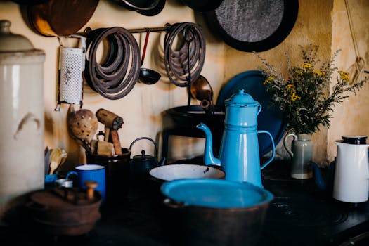 A cozy rustic kitchen features a vintage blue kettle with hanging pans and wildflowers, exuding warmth and charm.