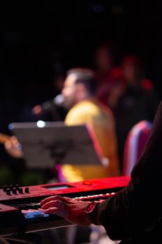 A close-up of a musician playing a keyboard during a live concert with a blurred band member in the background.