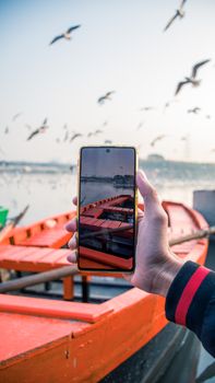 A hand holding a smartphone capturing birds flying over a lake with boats in the background.