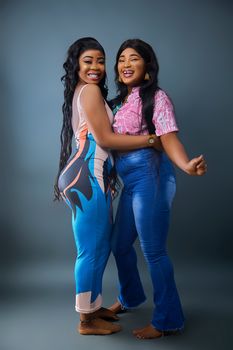 Two smiling women with long hair joyfully embracing in stylish outfits, showcasing friendship and happiness.