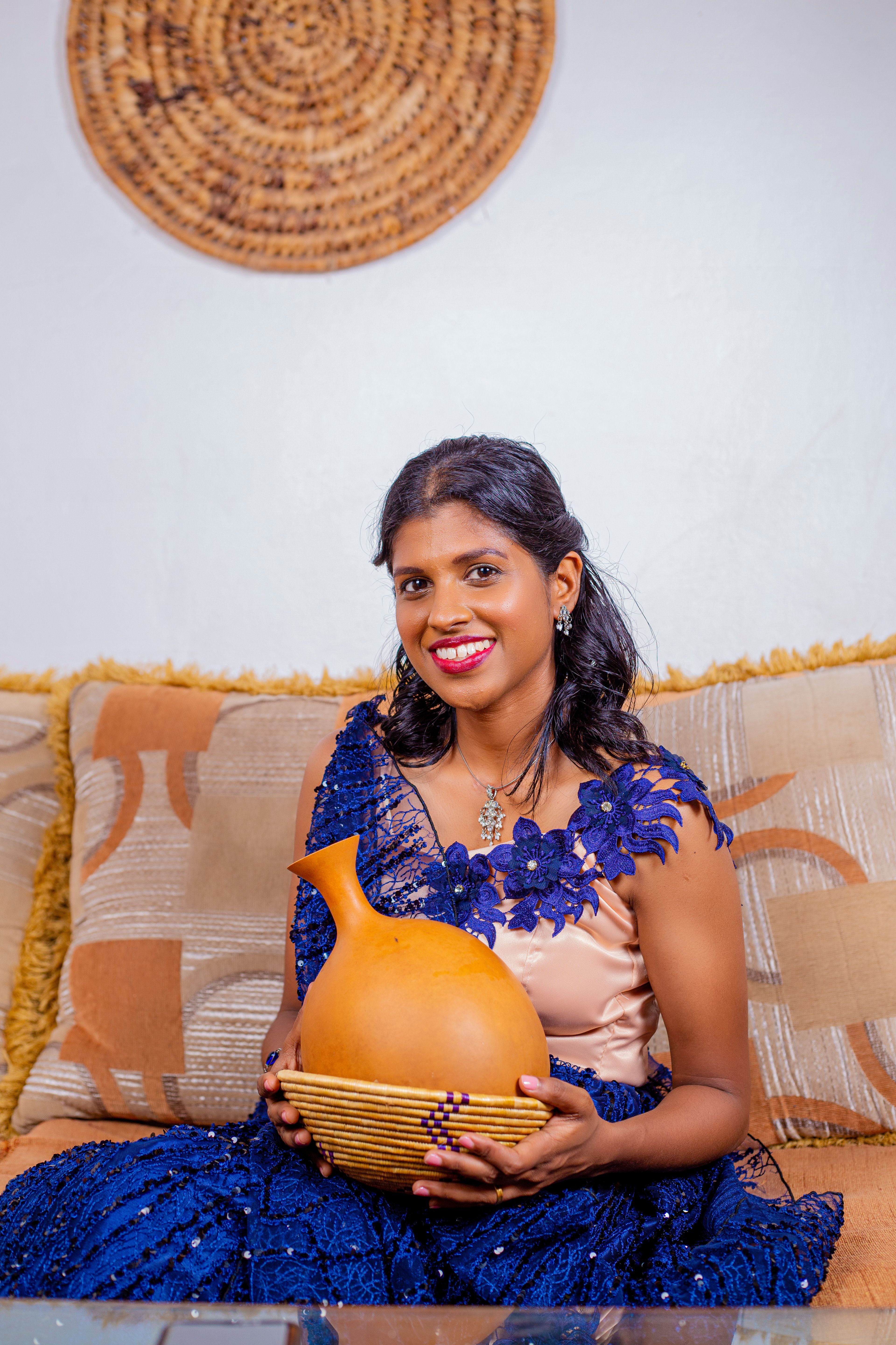 Smiling woman in blue gown holding a clay pot while sitting on a sofa, creating a warm and inviting atmosphere.