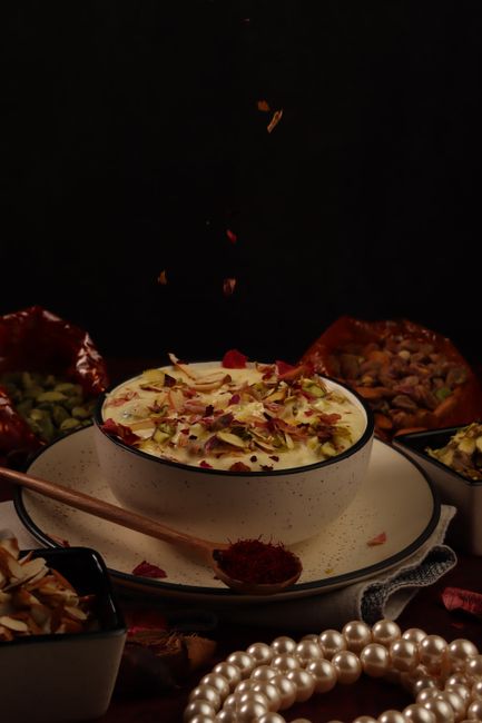Flattened white chenna discs floating in pale golden, thickened milk topped with slivered pistachios and saffron threads in a shallow bowl.