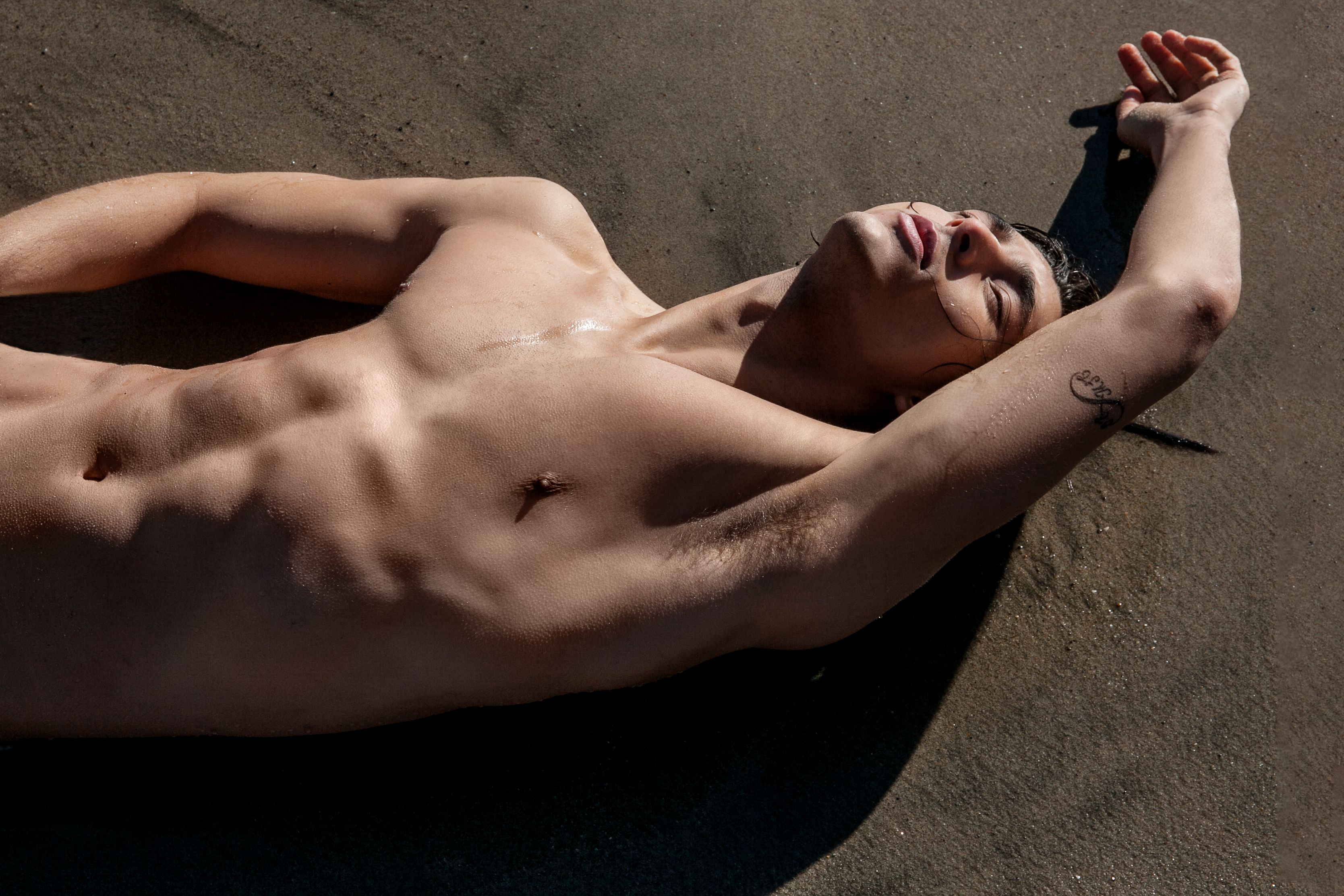 A muscular man sunbathing shirtless on the sandy beach with a tattoo visible.