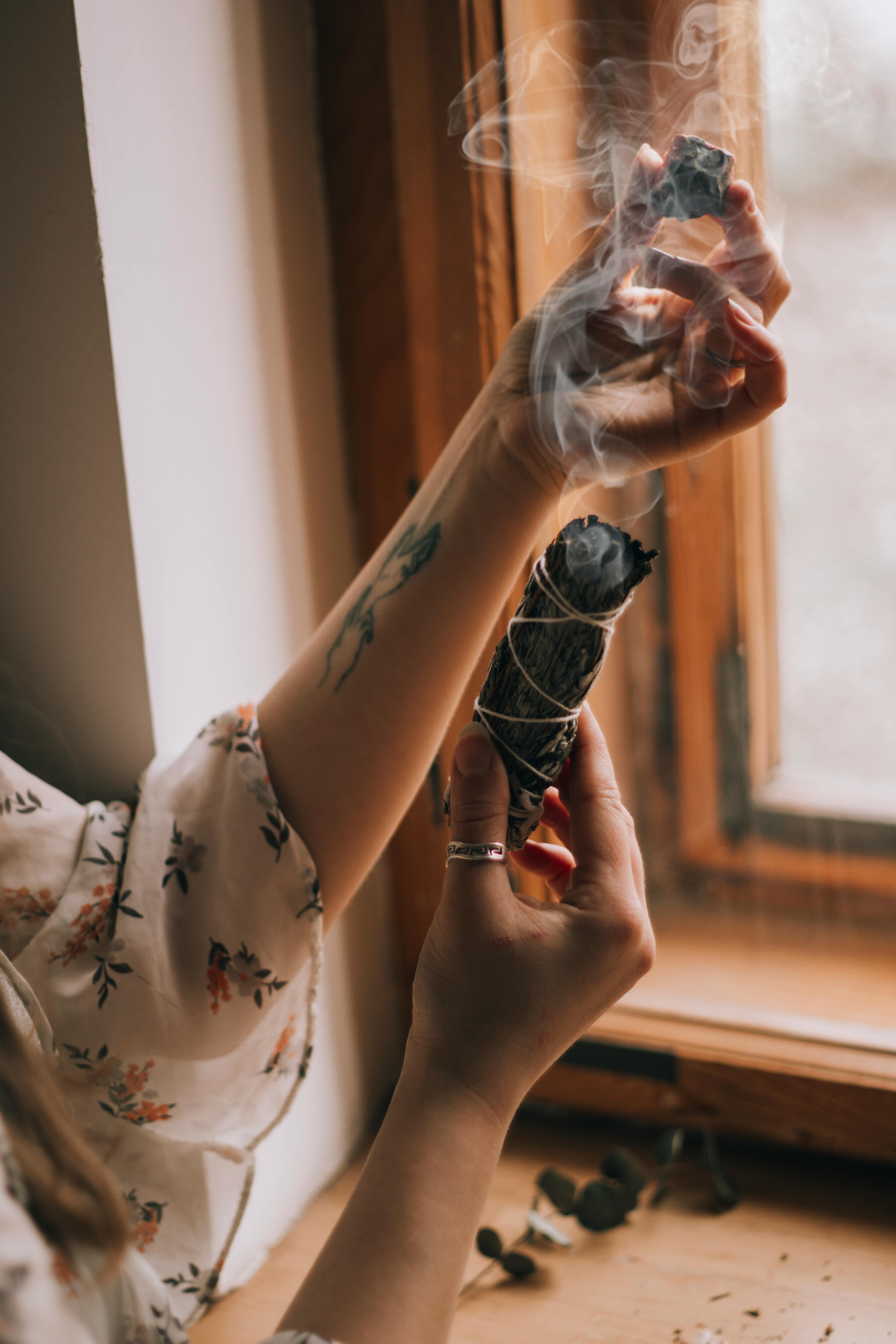 A calming scene of a woman using a smudge stick for aromatherapy near a window.