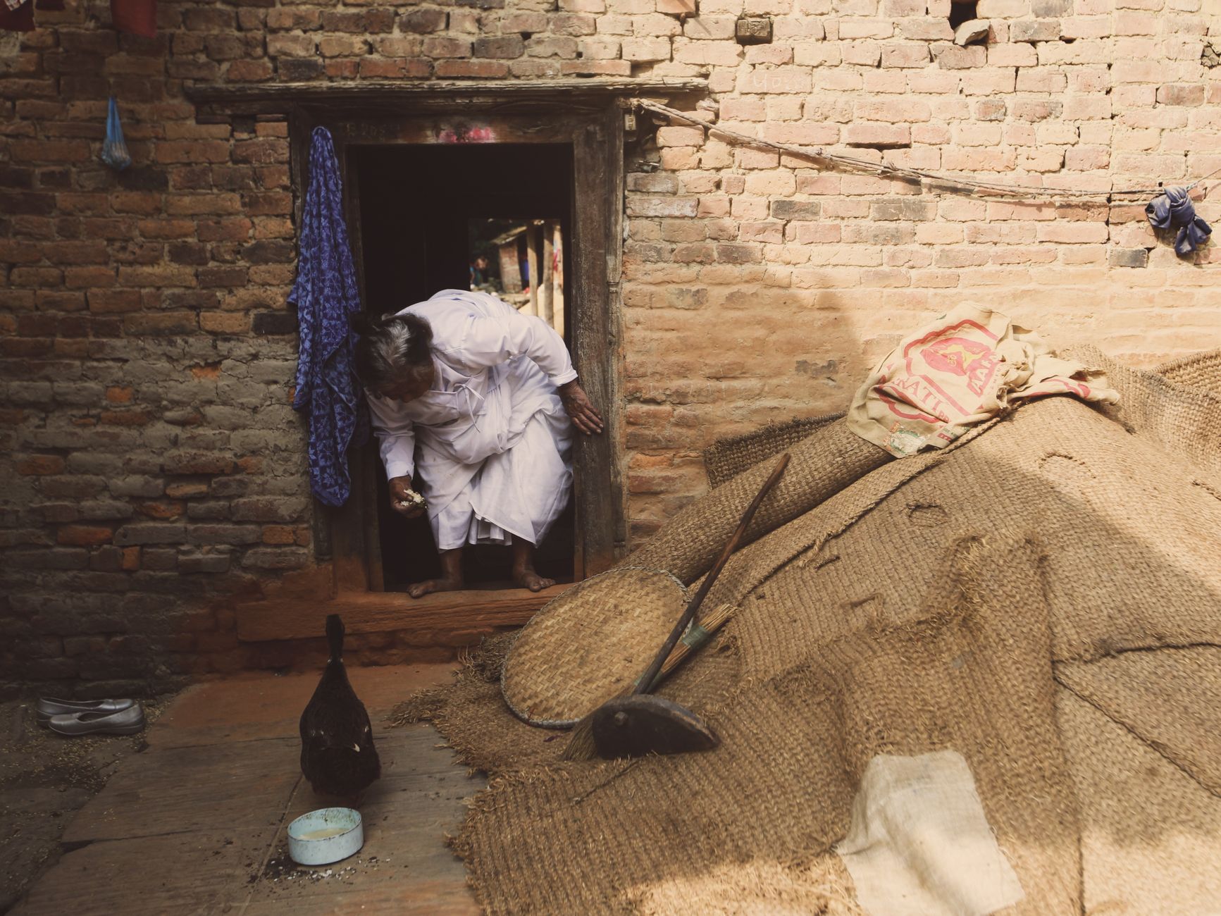 An elderly woman in traditional attire steps out of a rustic brick home. Urban setting.