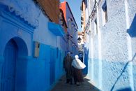 A couple walks through the vibrant blue-painted alleyways of Chefchaouen, Morocco, under the midday sun.