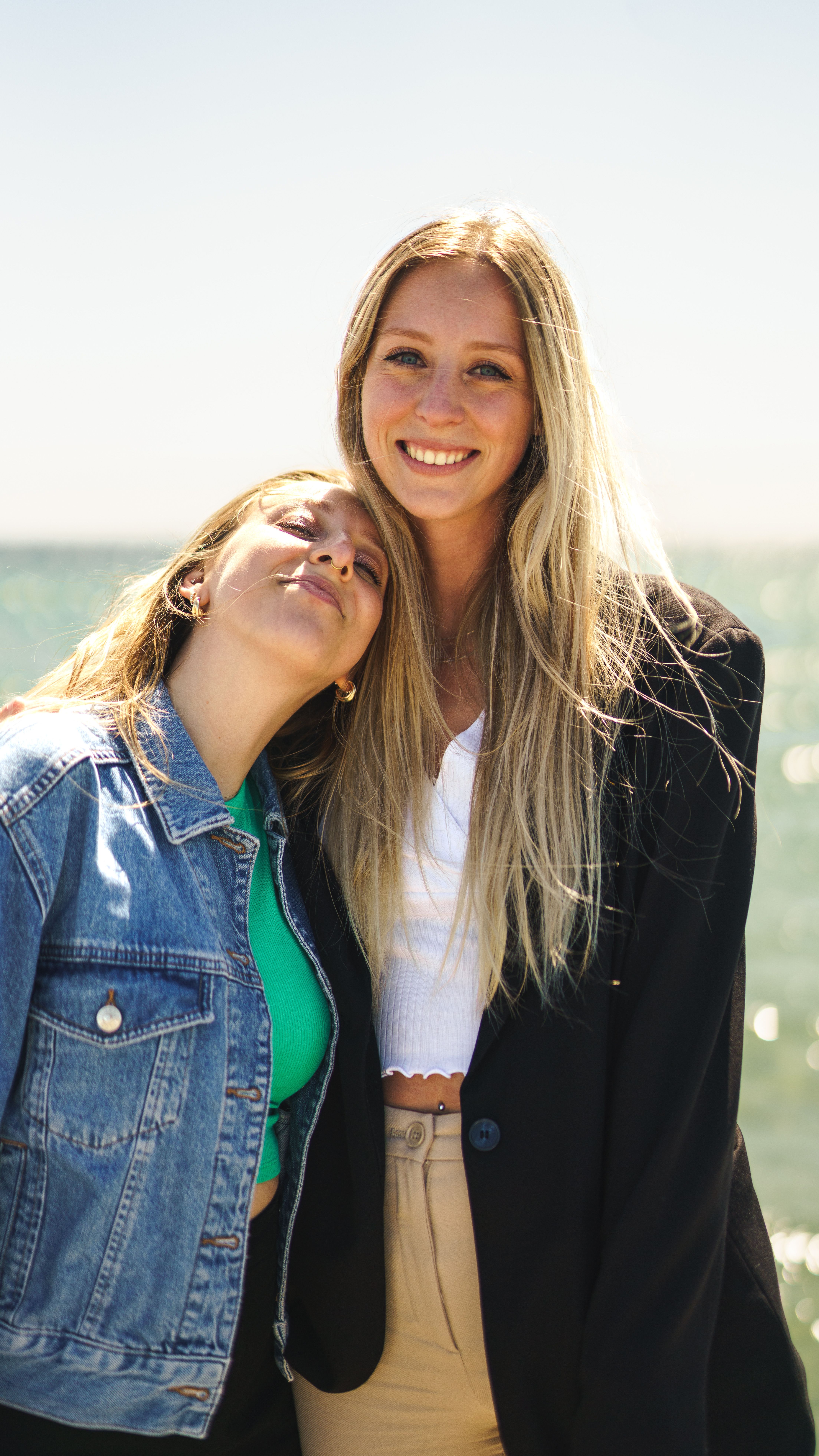 Two blonde women smile warmly near the ocean on a sunny day, exuding friendship and joy.