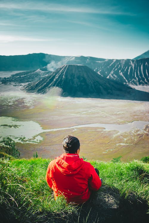 Back View of a Man in Red Hoodie Sitting on the Grass in Front of the Volcano