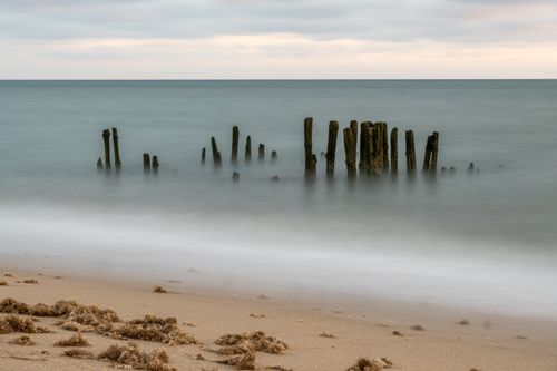 Free A long exposure captures a tranquil beach scene with remnants of old wooden poles emerging from the sea. Stock Photo