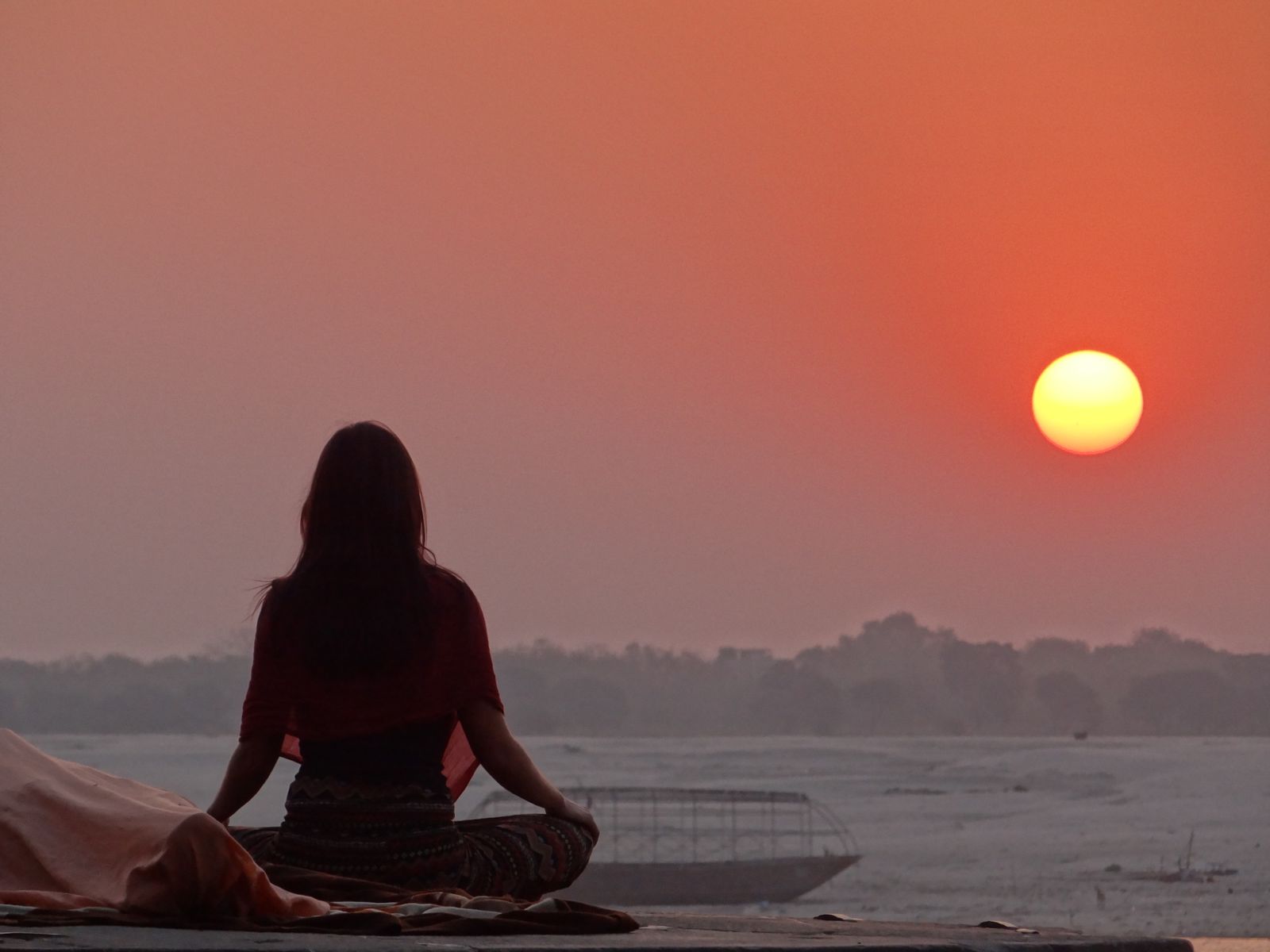 Woman meditating in disciplined stillness by a riverbank at sunrise, reflecting the practice of tapas