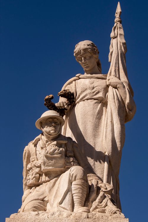 Free A low-angle view of a historical stone statue of a kneeling soldier with an allegorical figure under a clear blue sky. Stock Photo