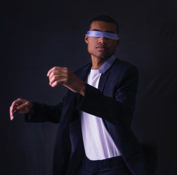 Portrait of a blindfolded man in a black suit, posing in a studio with a dark background.
