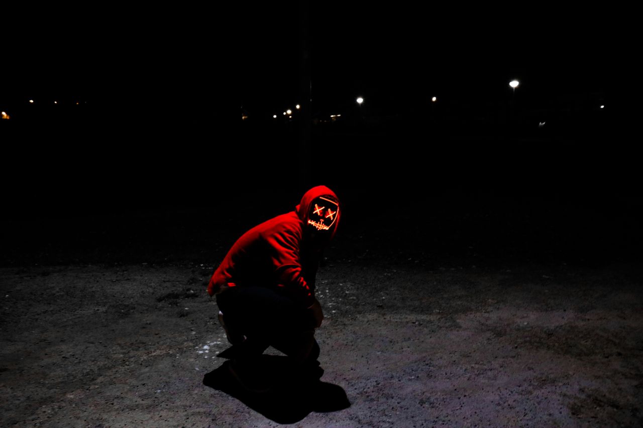 a person in a red hoodie with a glowing mask crouches in the shadows o