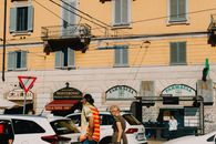 People walking in front of a pharmacy on a sunny urban street with traffic and shop signs.
