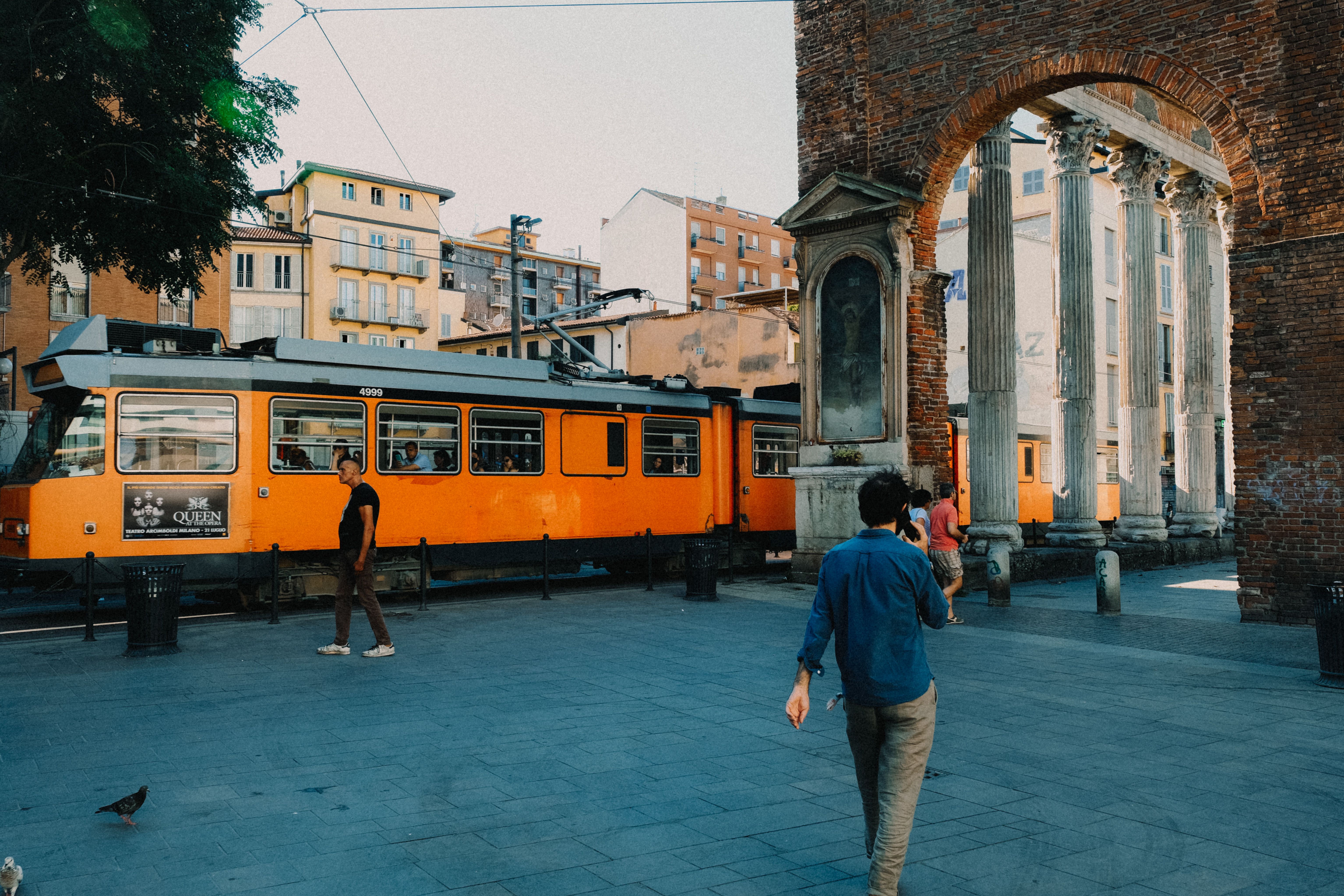 Vibrant urban scene featuring a tram passing by ancient architectural ruins and people walking nearby.