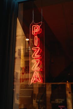 Vertical neon pizza sign illuminated in a dark window, creating a vintage urban vibe.