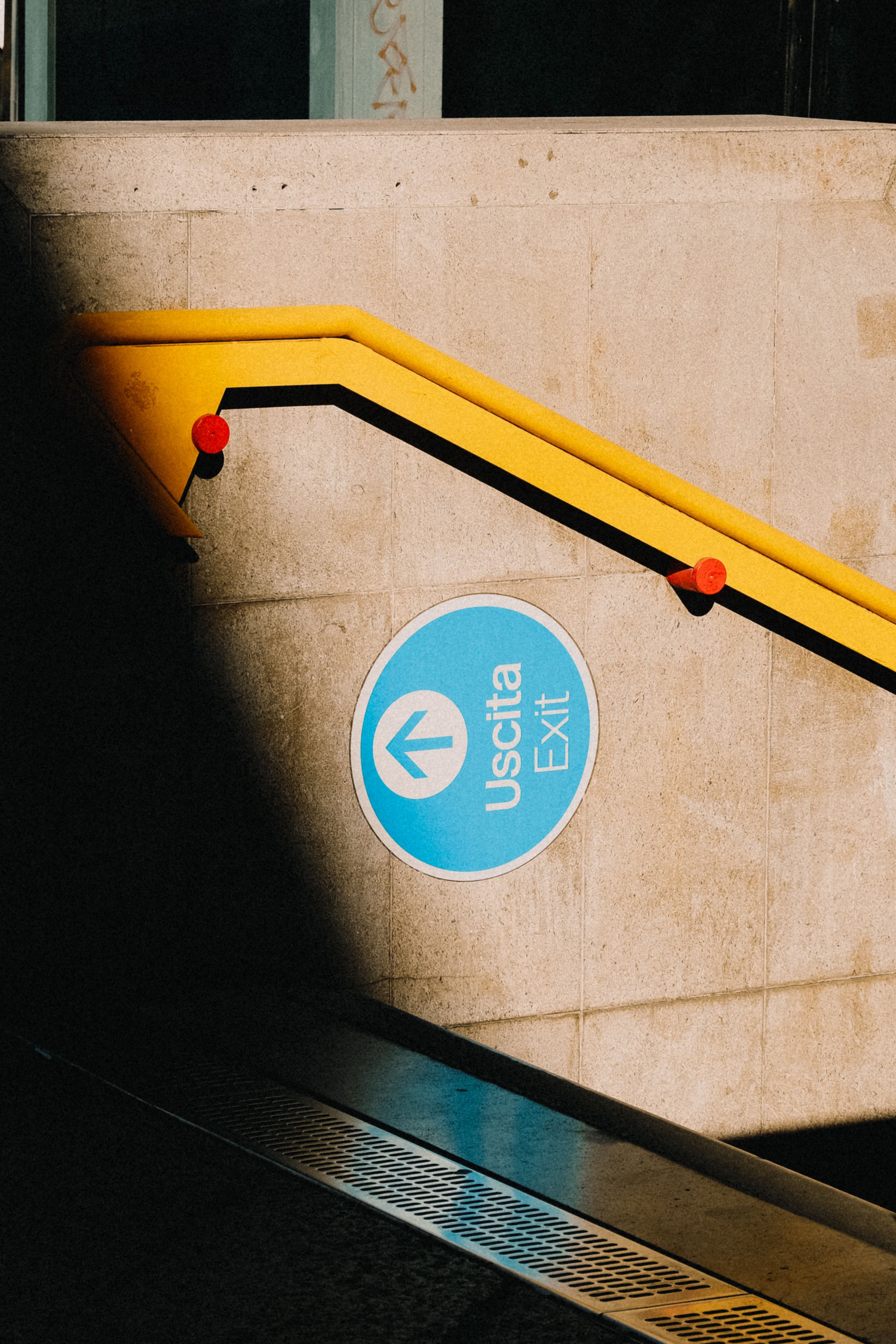 A blue 'Uscita Exit' sign with a yellow handrail in a subway station.