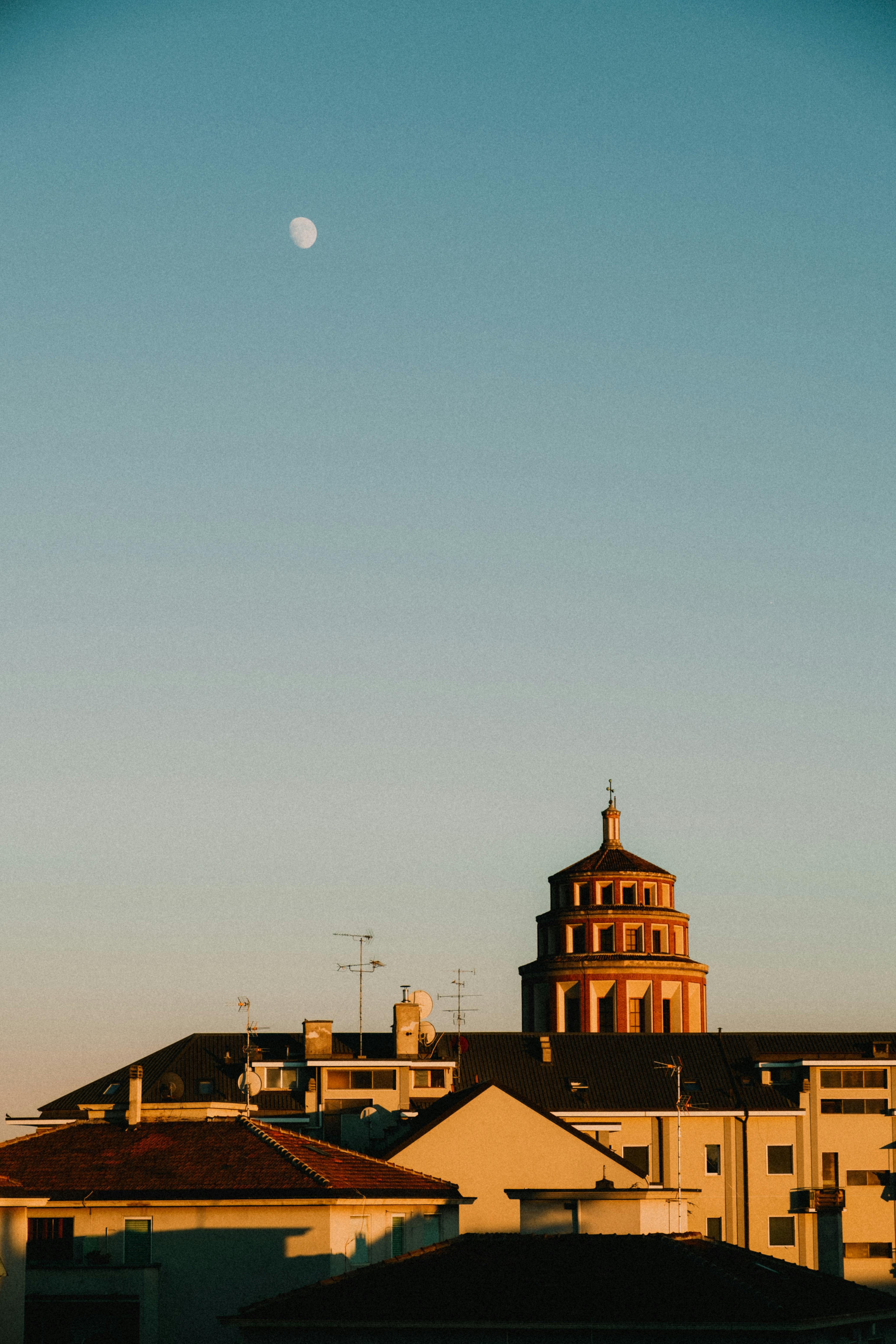 Cityscape at sunset featuring a historical building and the moon in a clear sky.