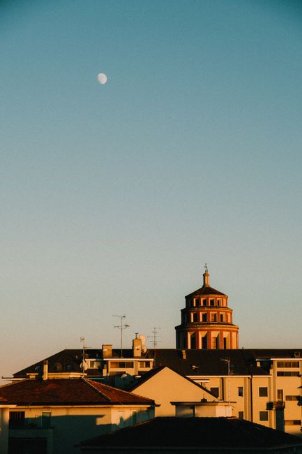Cityscape at sunset featuring a historical building and the moon in a clear sky.