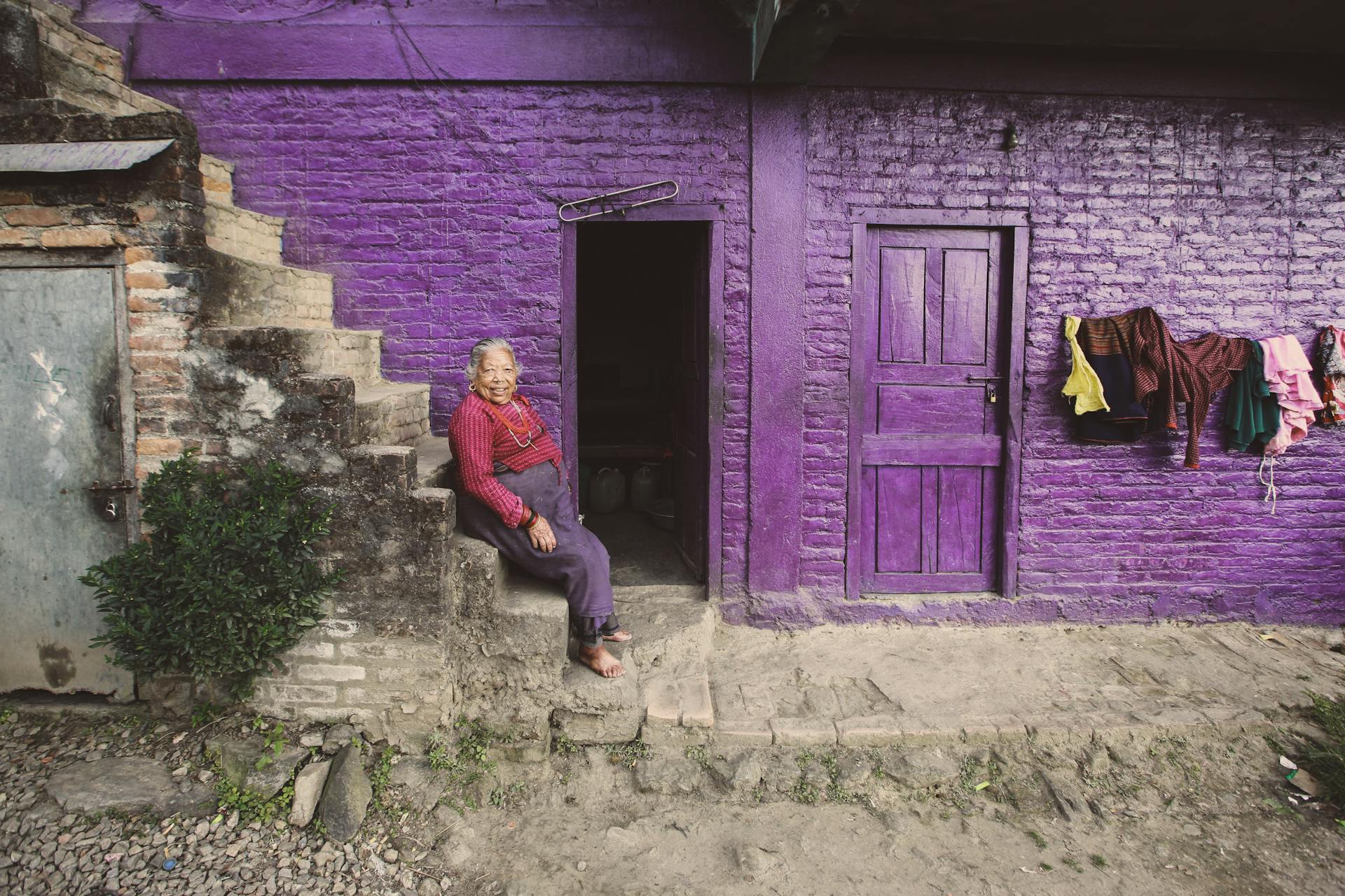 A senior woman sitting on steps of a purple-walled house with hanging laundry.