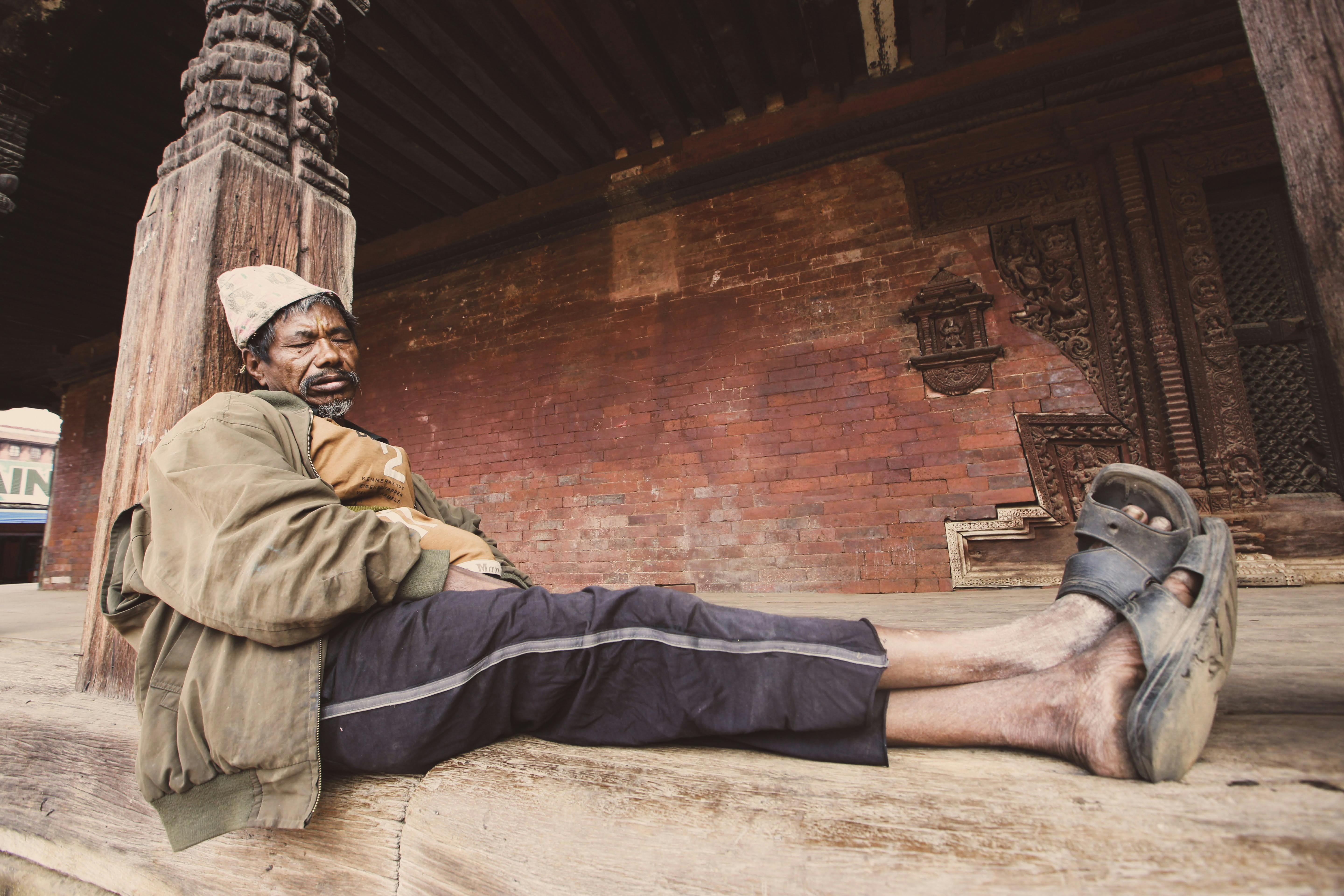 An elderly man rests against a historic brick wall, reflecting urban poverty.