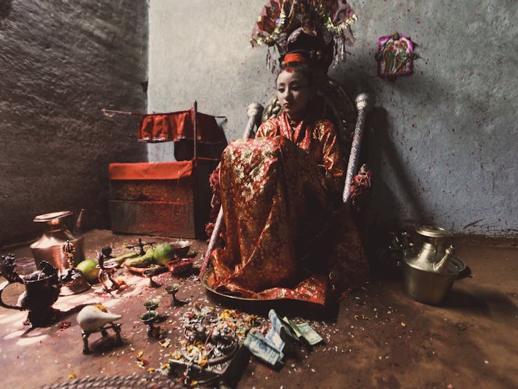 Young Girl Dressed As Deity Sitting Surrounded By Offerings